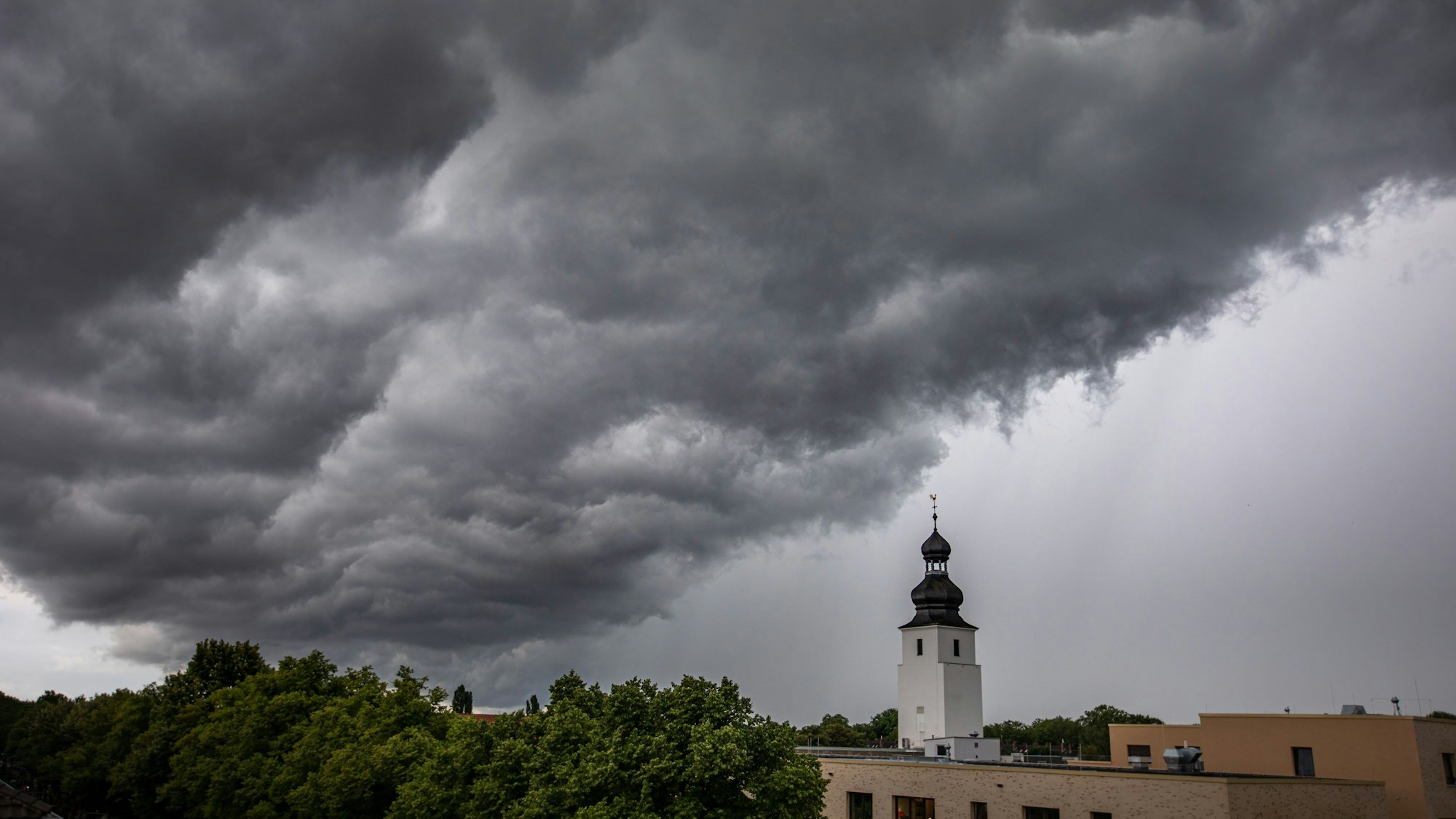 Dunkle Wolken ziehen am 4. Juli 2021 kurz vor einem Gewitter mit Starkregen über den Kölner Ortsteil Sülz. Der Kirchturm rechts gehört zur Kirche „Zur heiligen Familie“.
