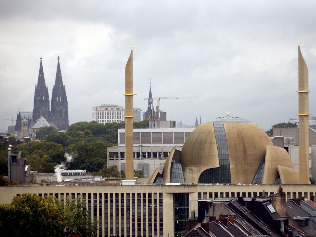 Im Vordergrund ragen zwei Minarette der Großmoschee in den Himmel, im Hintergrund steht der Kölner Dom.