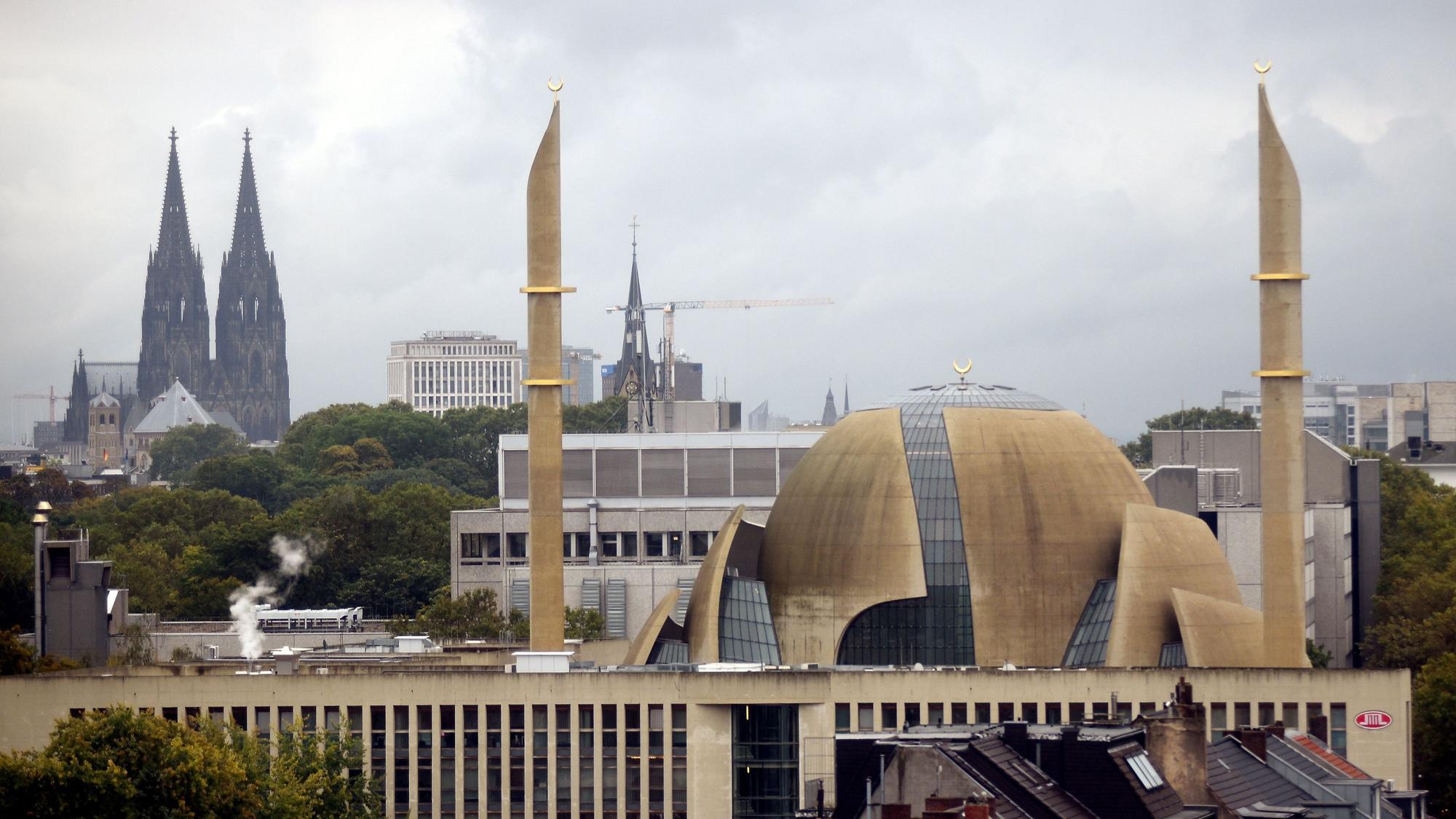 Im Vordergrund ragen zwei Minarette der Großmoschee in den Himmel, im Hintergrund steht der Kölner Dom.