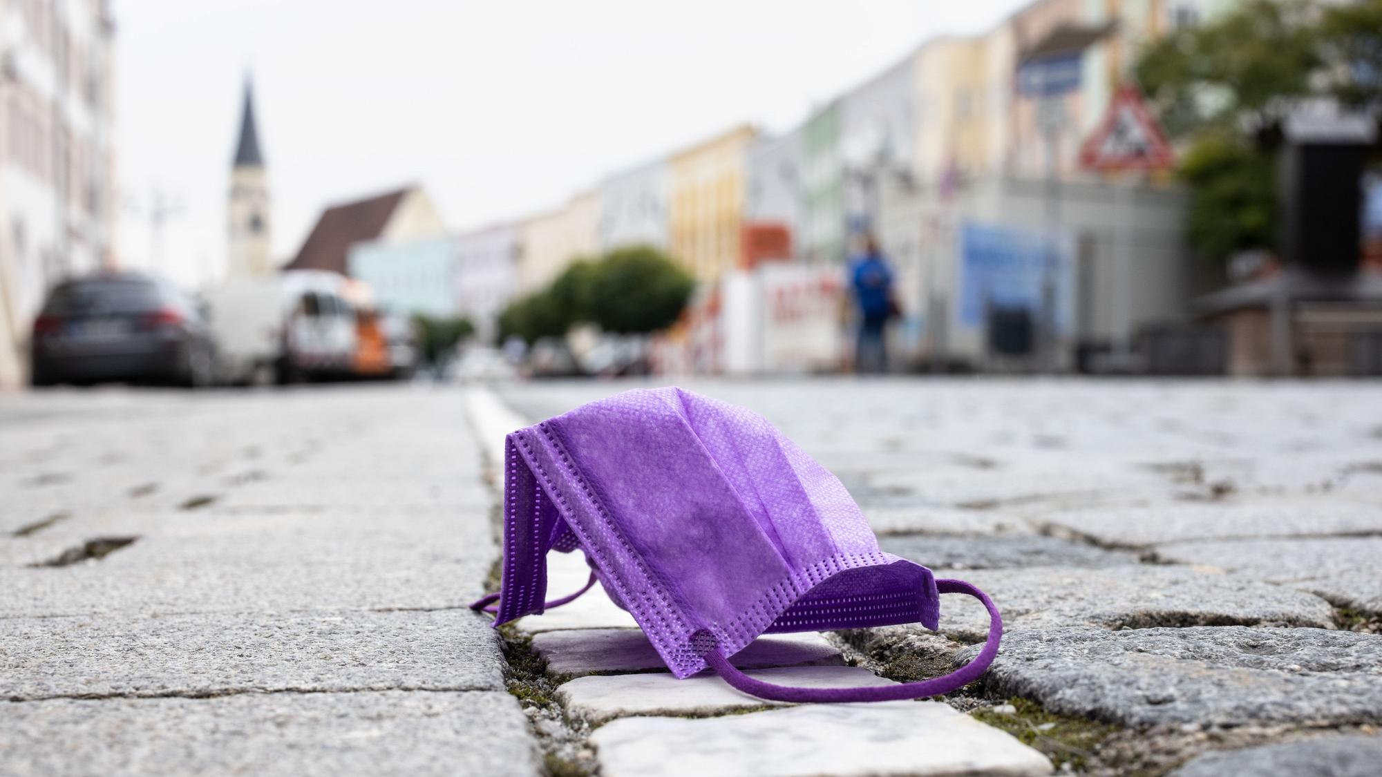 Bayern kämpft mit immer höher steigenden Corona-Zahlen. Auch die Bürger sind entzweit: Während die einen endlich lockerere Maßnahmen fordern, wollen die anderen härtere Maßnahmen, um die Infektionswelle einzudämmen. Auf dem Foto (aufgenommen am 26. Oktober 2021) zu sehen: Eine violette medizinische Maske, die auf einer Straße liegt, während im Hintergrund die Altstadt von Mühldorf am Inn zu sehen ist.