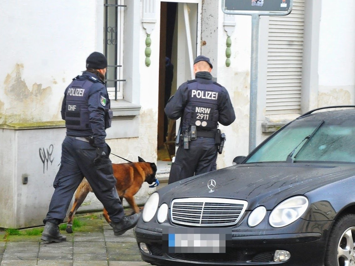 Polizisten und ein Spürhund stehen vor einem Haus.
