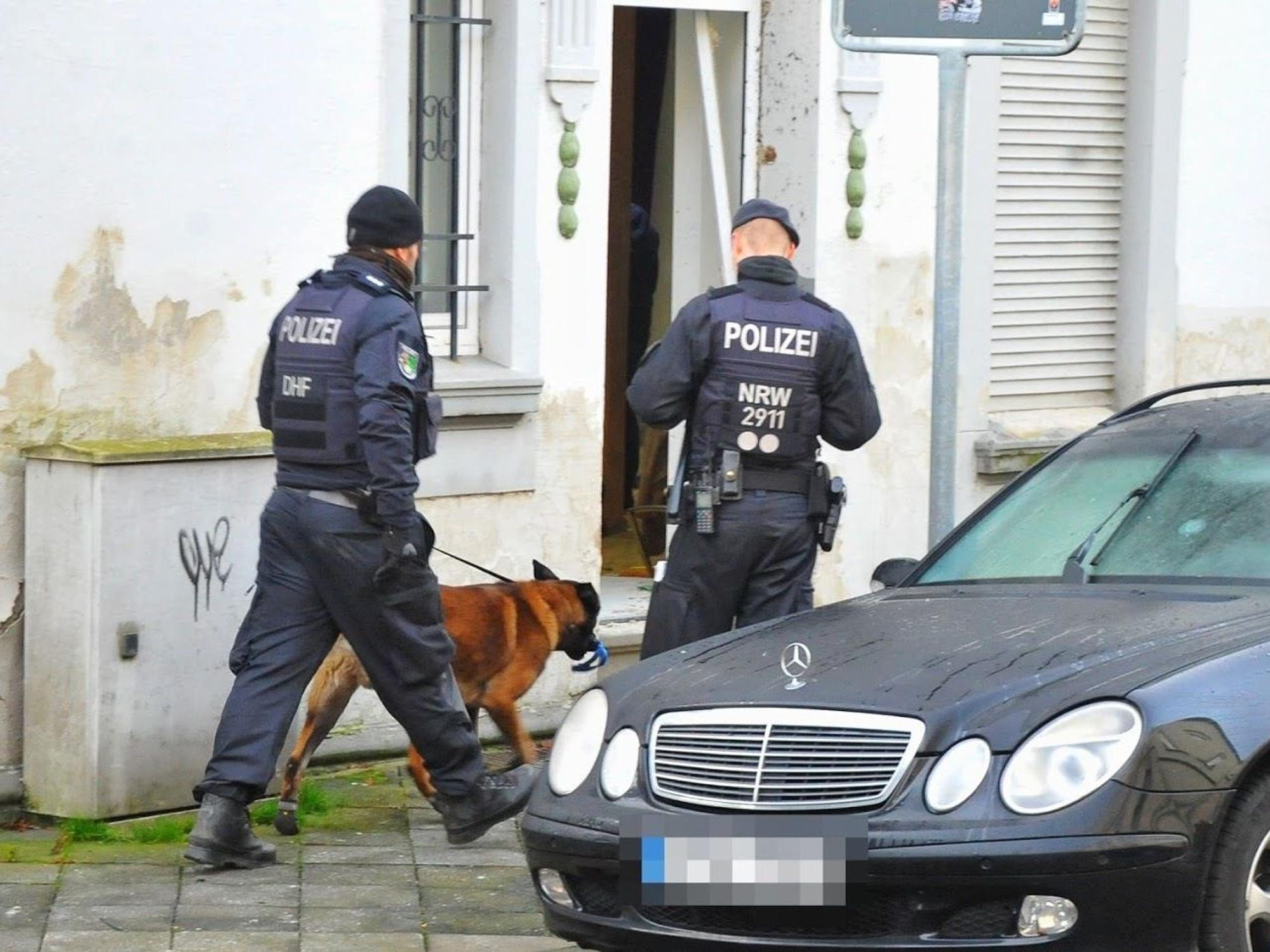 Polizisten und ein Spürhund stehen vor einem Haus.