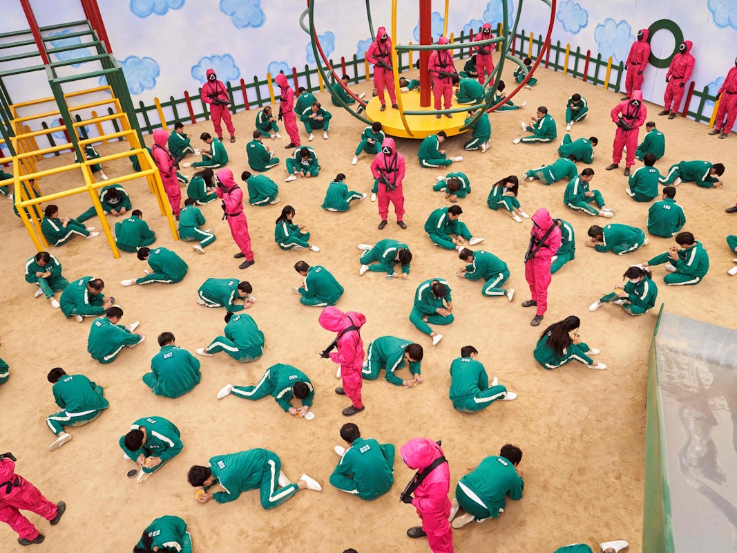 Szene aus der Netflix-Serie „Squid Game“, in der hunderte Erwachsene in Kinderspielen gegeneinander antreten. Die Teilnehmer sitzen auf dem Boden eines nachgebauten Spielplatzes und schnitzen Salzformen aus.