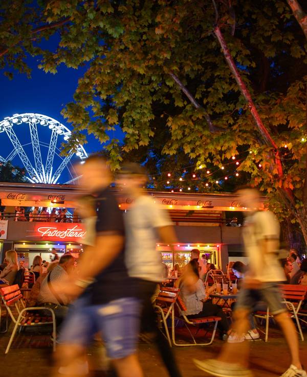 Passanten laufen in unserem Symbolbild im Juni 2021 an einem Sommerabend am Erzsébet Platz vor dem „Budapest Eye“-Riesenrad im Stadtteil Pest an Bars und Außengastronomie entlang.