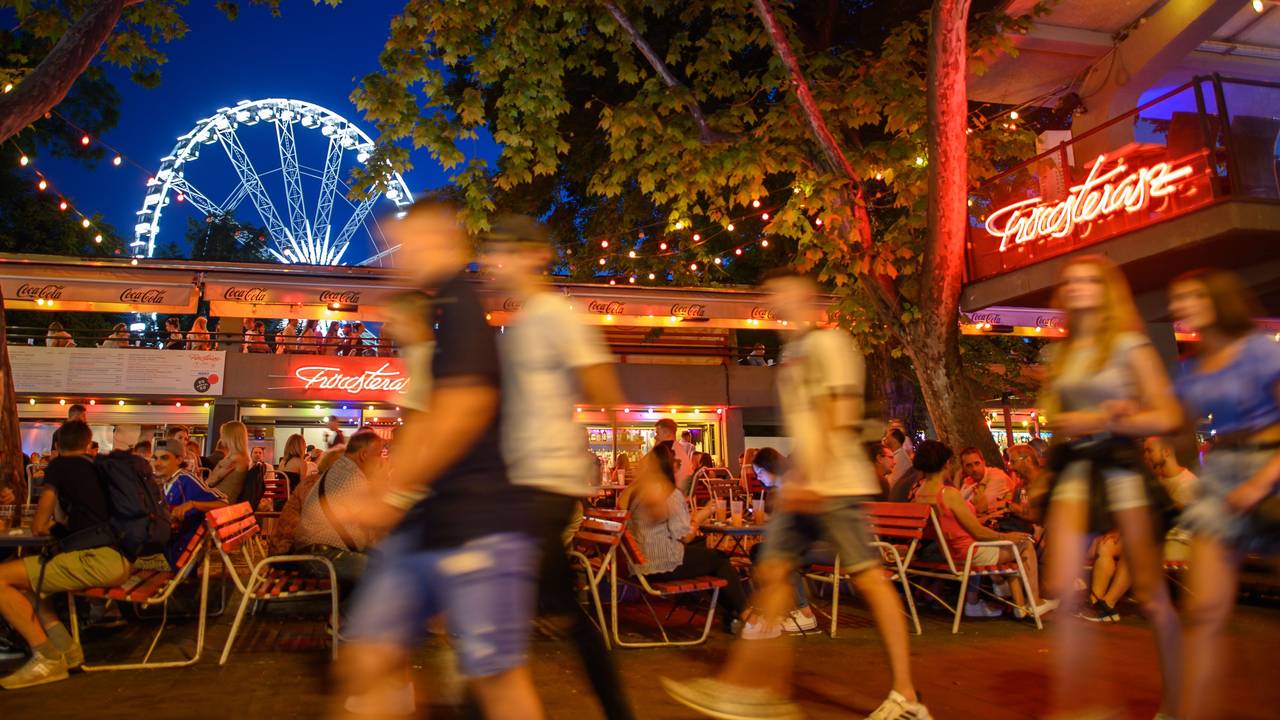 Passanten laufen in unserem Symbolbild im Juni 2021 an einem Sommerabend am Erzsébet Platz vor dem „Budapest Eye“-Riesenrad im Stadtteil Pest an Bars und Außengastronomie entlang.