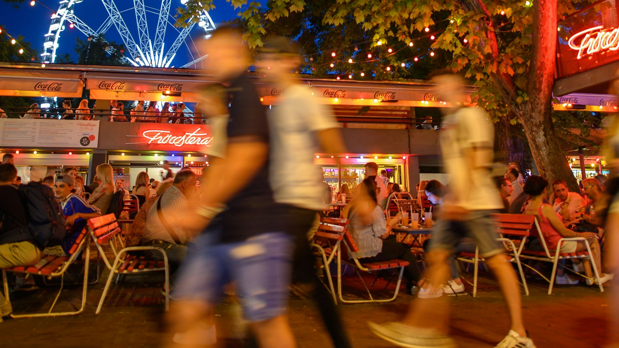 Passanten laufen in unserem Symbolbild im Juni 2021 an einem Sommerabend am Erzsébet Platz vor dem „Budapest Eye“-Riesenrad im Stadtteil Pest an Bars und Außengastronomie entlang.