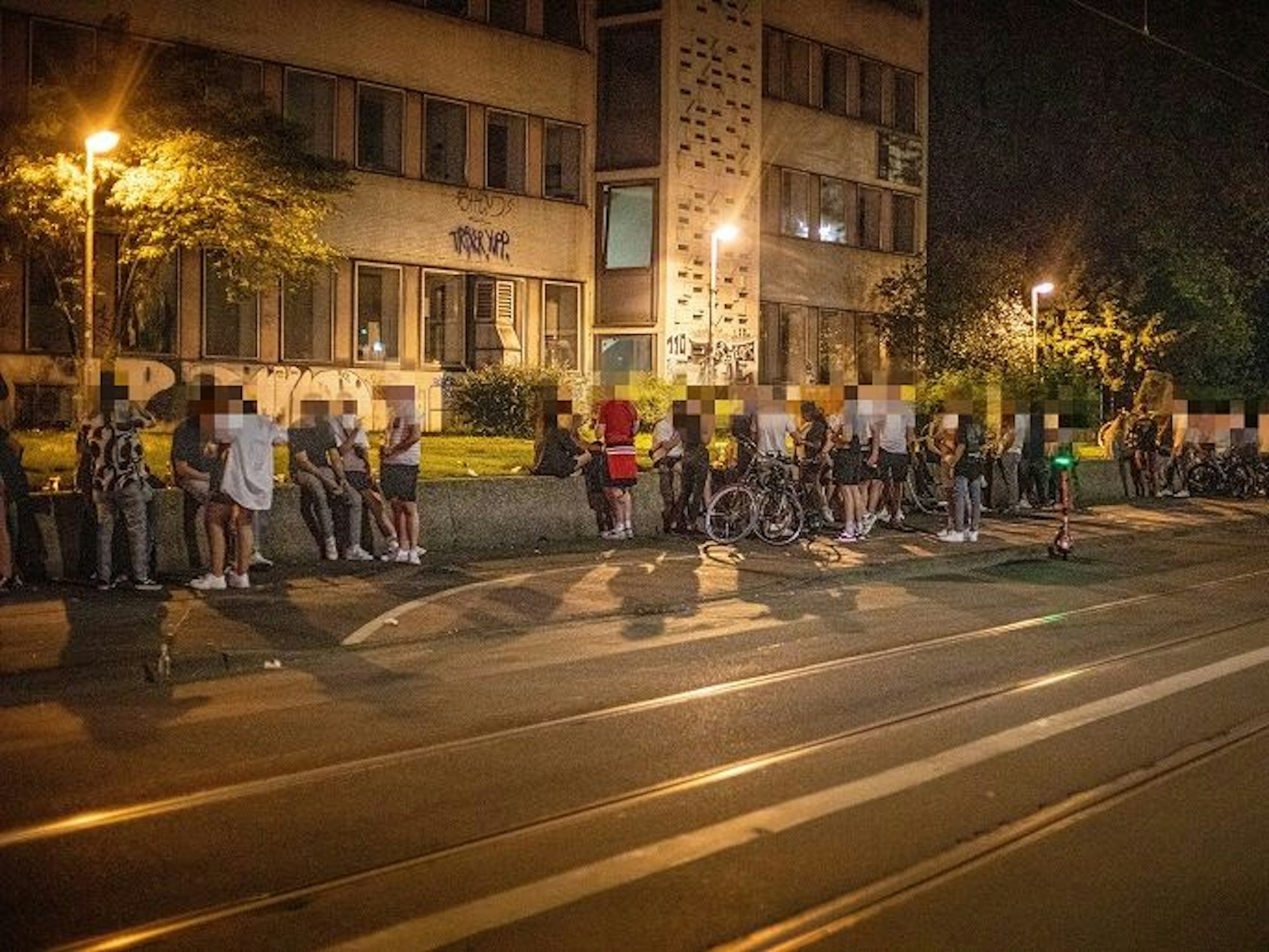 Auf einer Mauer an der Zülpicher Straße auf dem Universitätsgelände sitzen zahlreiche Menschen.
