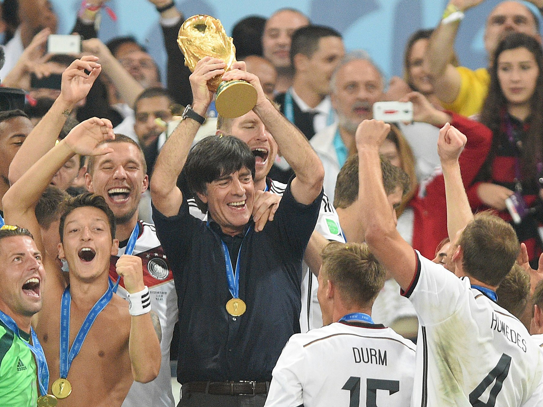 Joachim Löw hält den Weltmeisterpokal in den Händen. Um ihn tummeln sich seine Spieler. Unter anderem Roman Weidenfeller, Mesut Özil, Lukas Podolski, Per Mertesacker, Erik Durm und Benedikt Höwedes.
