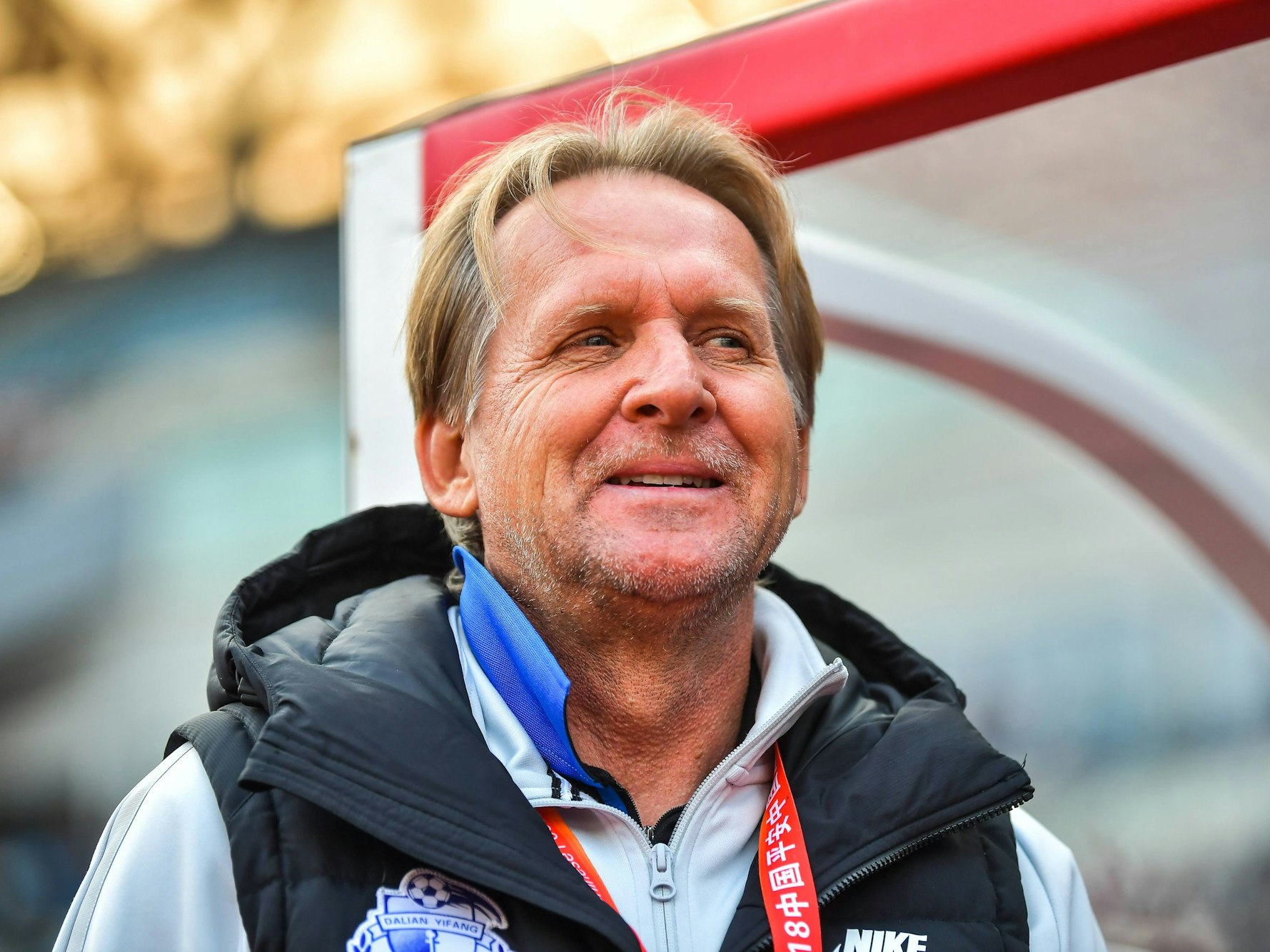 Bernd Schuster steht vor der Trainerbank und lächelt.