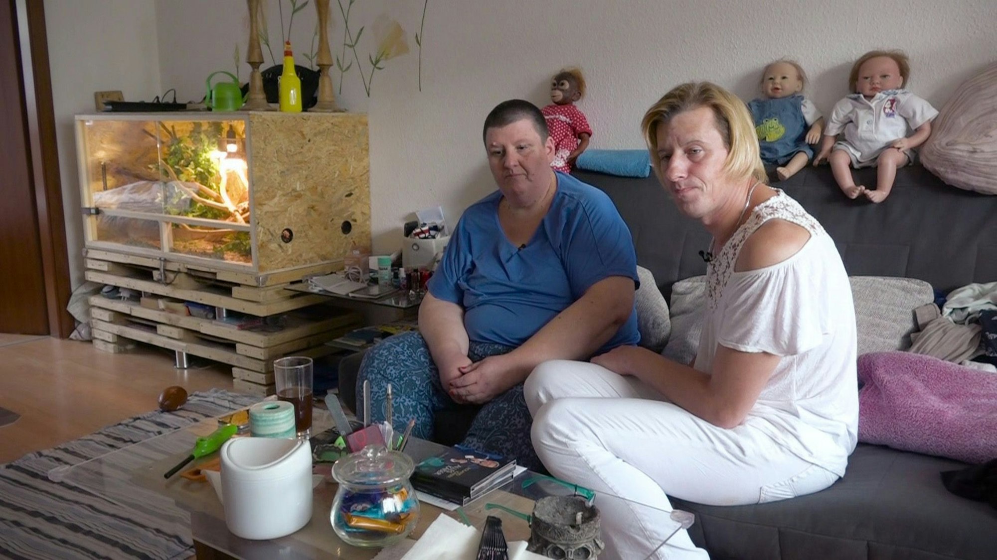 Sonja (r.) und Claudia (l.) sitzen auf einer Couch in ihrer Wohnung in Leverkusen. In der Ecke ist das Schlangenterrarium zu sehen.