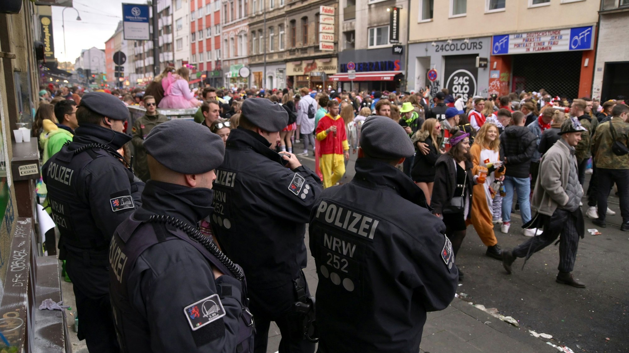 Feiernde Jecken ziehen durch eine Straße in Köln, am Rand stehen fünf Polizisten.