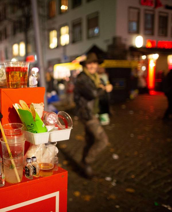 Abfall türmt sich auf einem Mülleimer. Karnevalisten feiern in den Straßen der Kölner Altstadt in den Abend hinein.