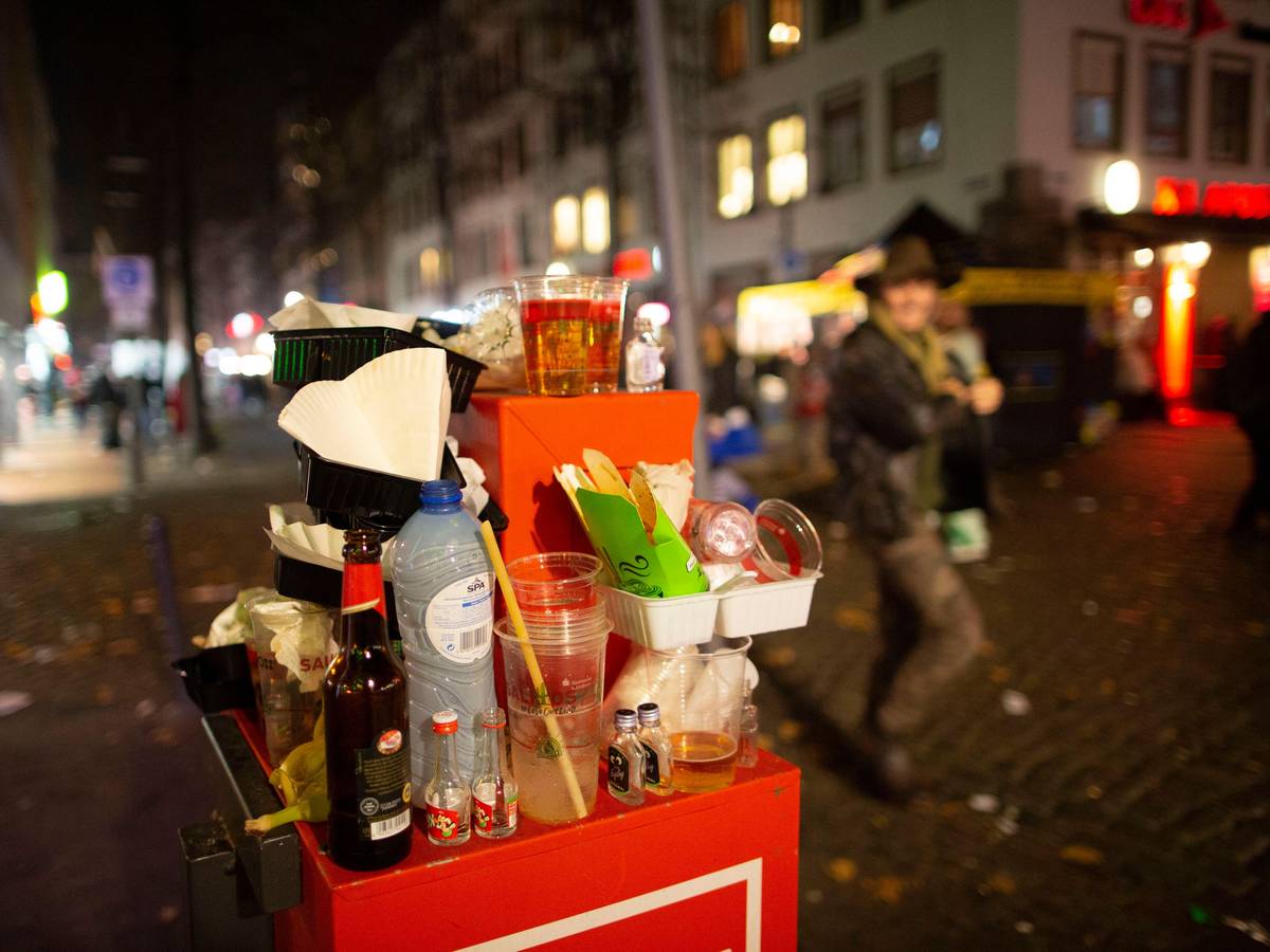 Abfall türmt sich auf einem Mülleimer. Karnevalisten feiern in den Straßen der Kölner Altstadt in den Abend hinein.