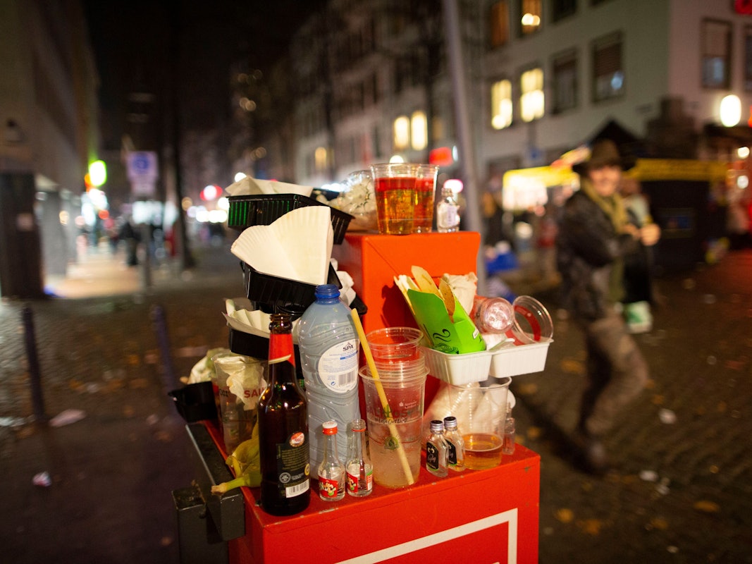 Abfall türmt sich auf einem Mülleimer. Karnevalisten feiern in den Straßen der Kölner Altstadt in den Abend hinein.