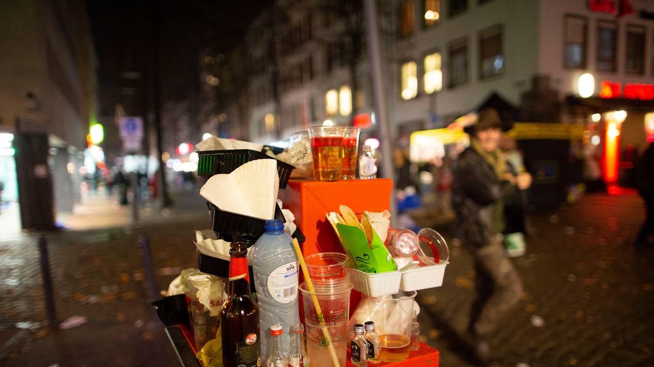 Abfall türmt sich auf einem Mülleimer. Karnevalisten feiern in den Straßen der Kölner Altstadt in den Abend hinein.