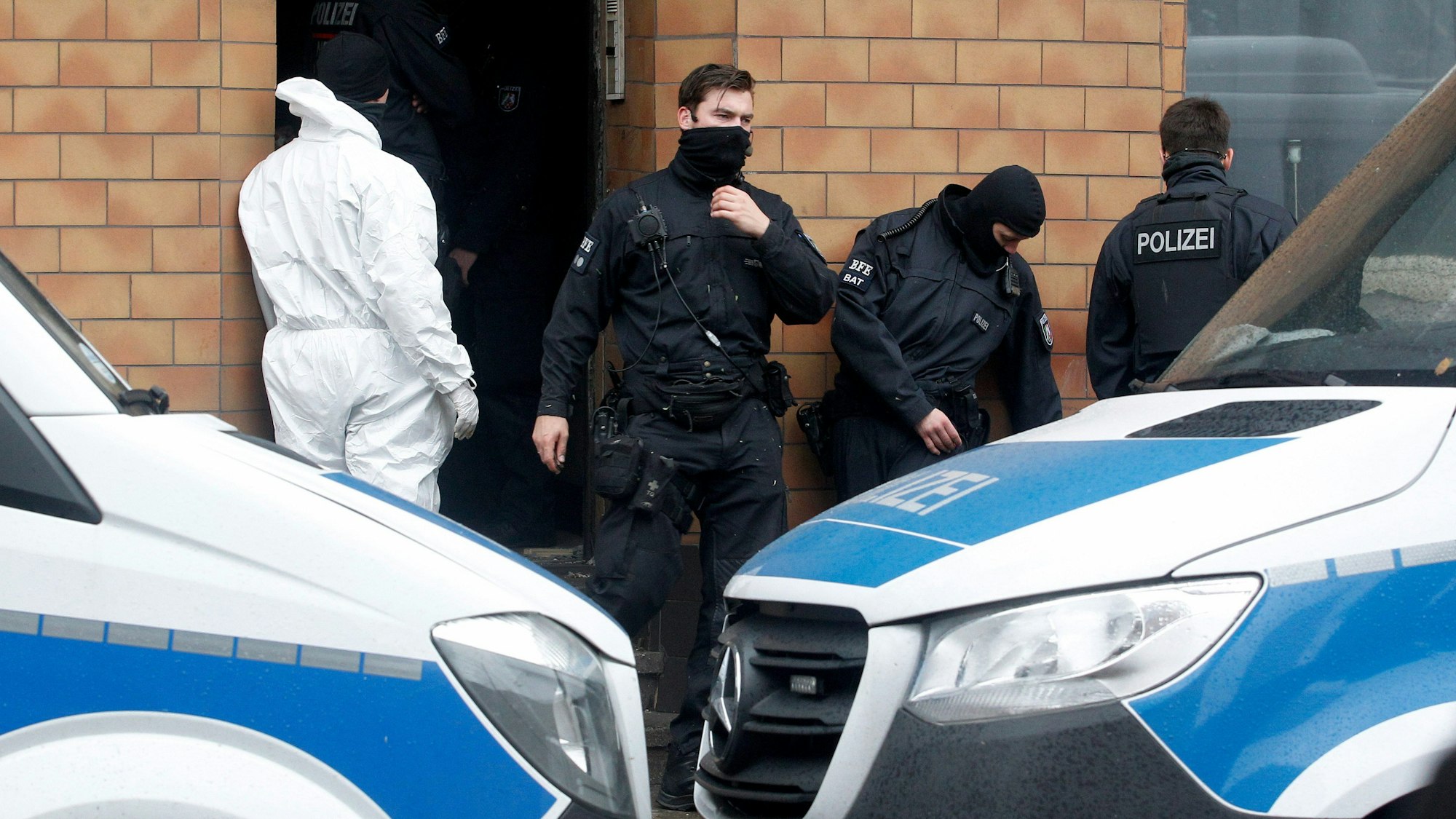 Vier Einsatzkräfte der Polizei, einer von der Spurensicherung im Schutzoverall, stehen vor einem Hauseingang. Davor parken zwei Polizeifahrzeuge.