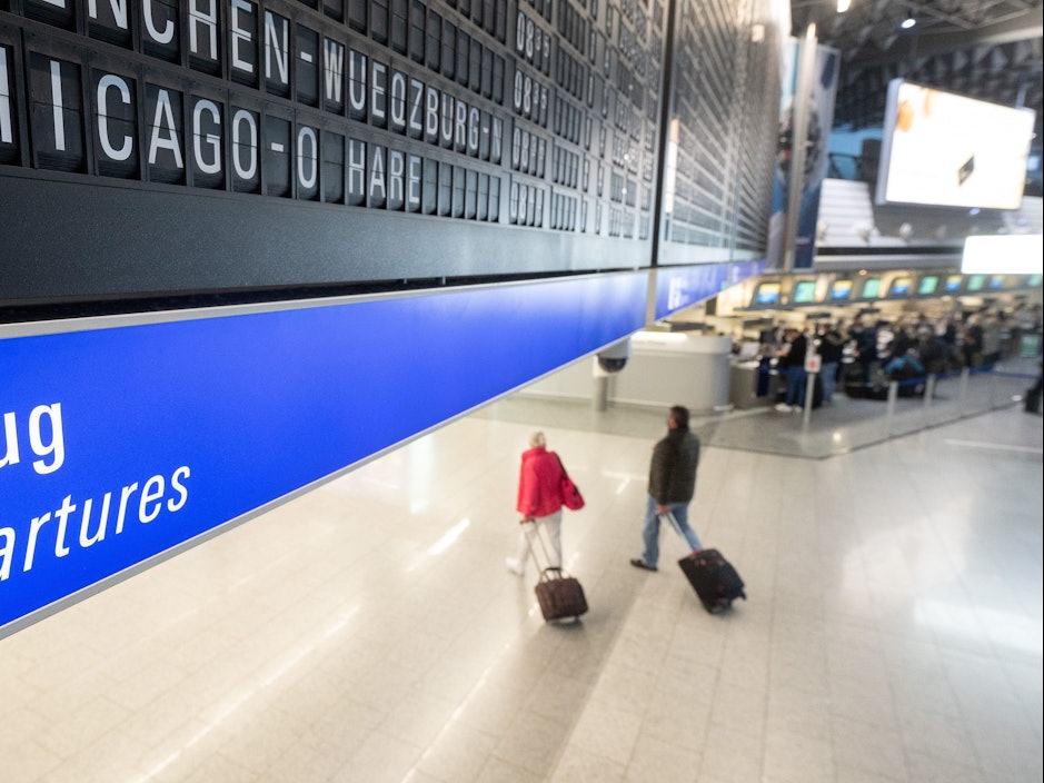 Ein Flug nach Chicago O·Hare in den USA wird auf dem Flughafen Frankfurt auf einer Tafel angezeigt unter der Passagiere mit Koffern entlang gehen.