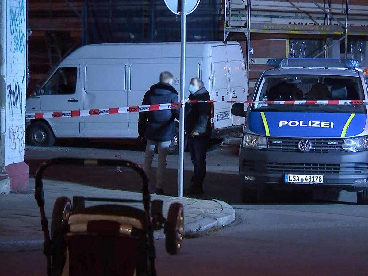 Ein umgestürzter Kinderwagen liegt auf der Straße, während im Hintergrund Polizeibeamte vor einem weißen Transporter stehen.