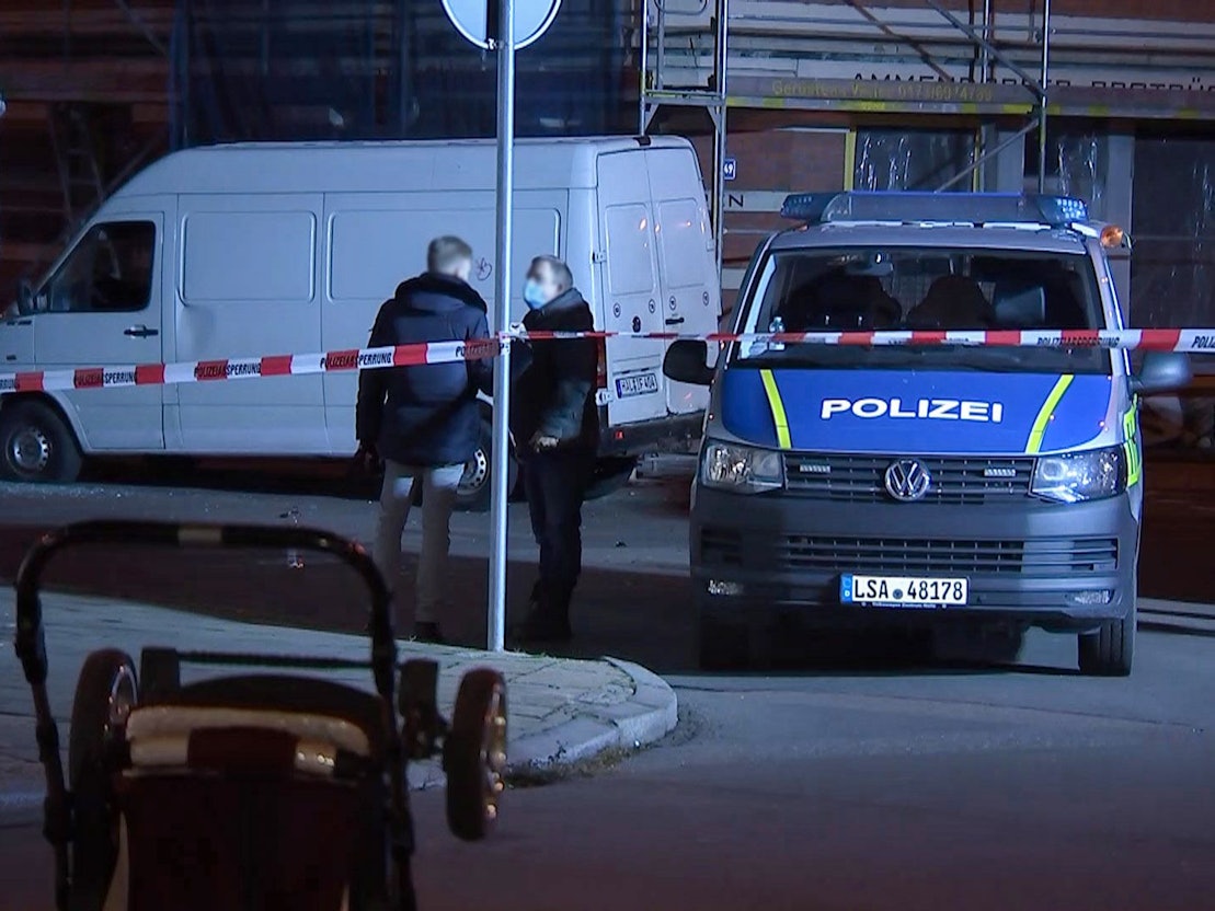 Ein umgestürzter Kinderwagen liegt auf der Straße, während im Hintergrund Polizeibeamte vor einem weißen Transporter stehen.