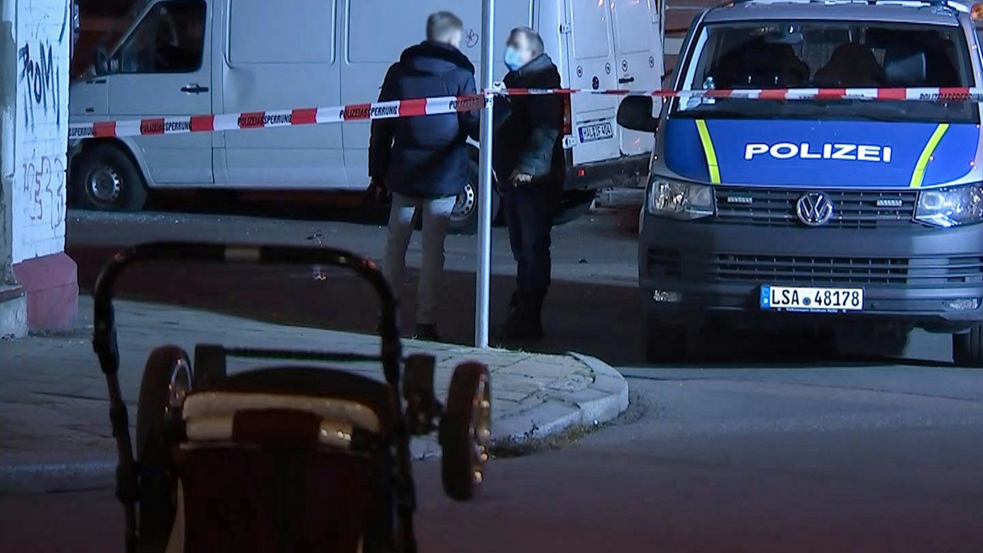 Ein umgestürzter Kinderwagen liegt auf der Straße, während im Hintergrund Polizeibeamte vor einem weißen Transporter stehen.