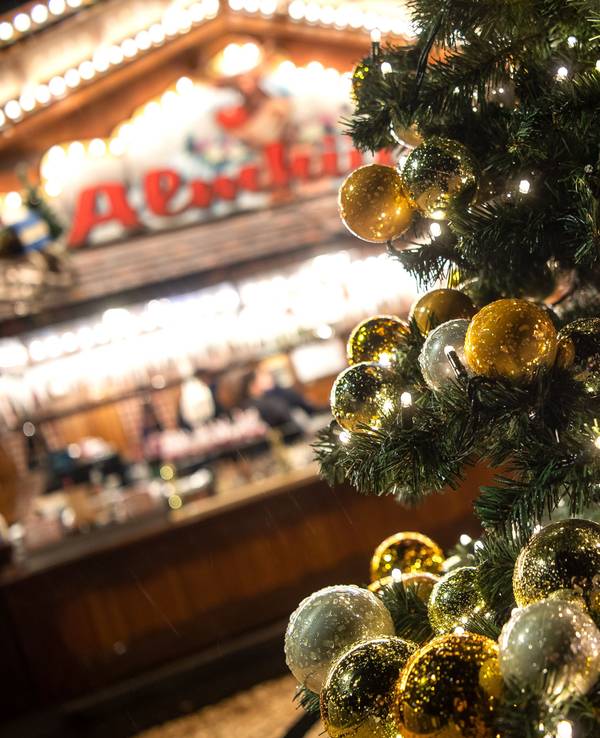 Auf dem Symbolfoto sieht man einen geschmückten Weihnachtsbaum, der vor einem beleuchteten Weihnachtsstand leuchtet.