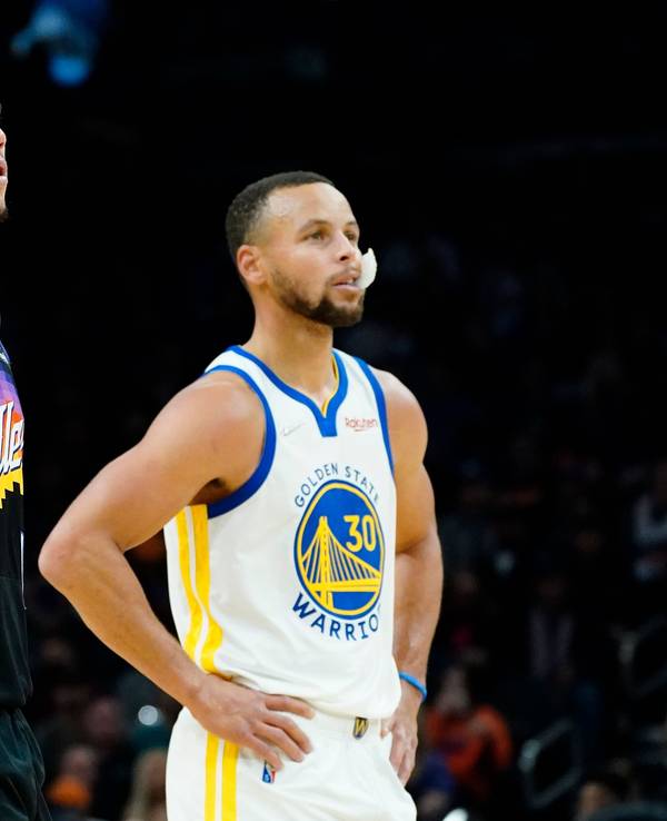 Suns-Guard Devin Booker (l.) und Warriors-Star Stephen Curry (r.) stützen jeweils ihre Arme in die Hüfte und schauen in Richtung Korb.