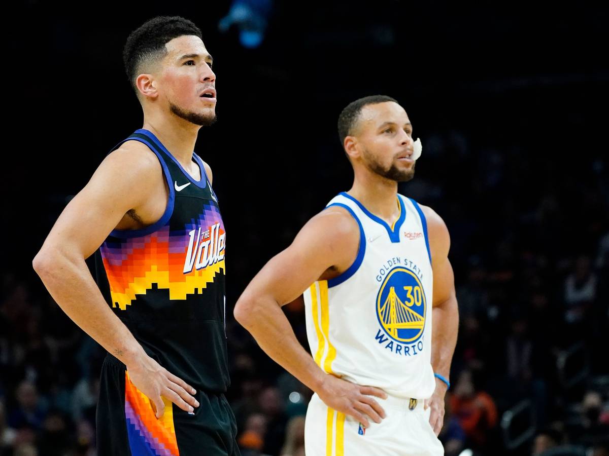 Suns-Guard Devin Booker (l.) und Warriors-Star Stephen Curry (r.) stützen jeweils ihre Arme in die Hüfte und schauen in Richtung Korb.