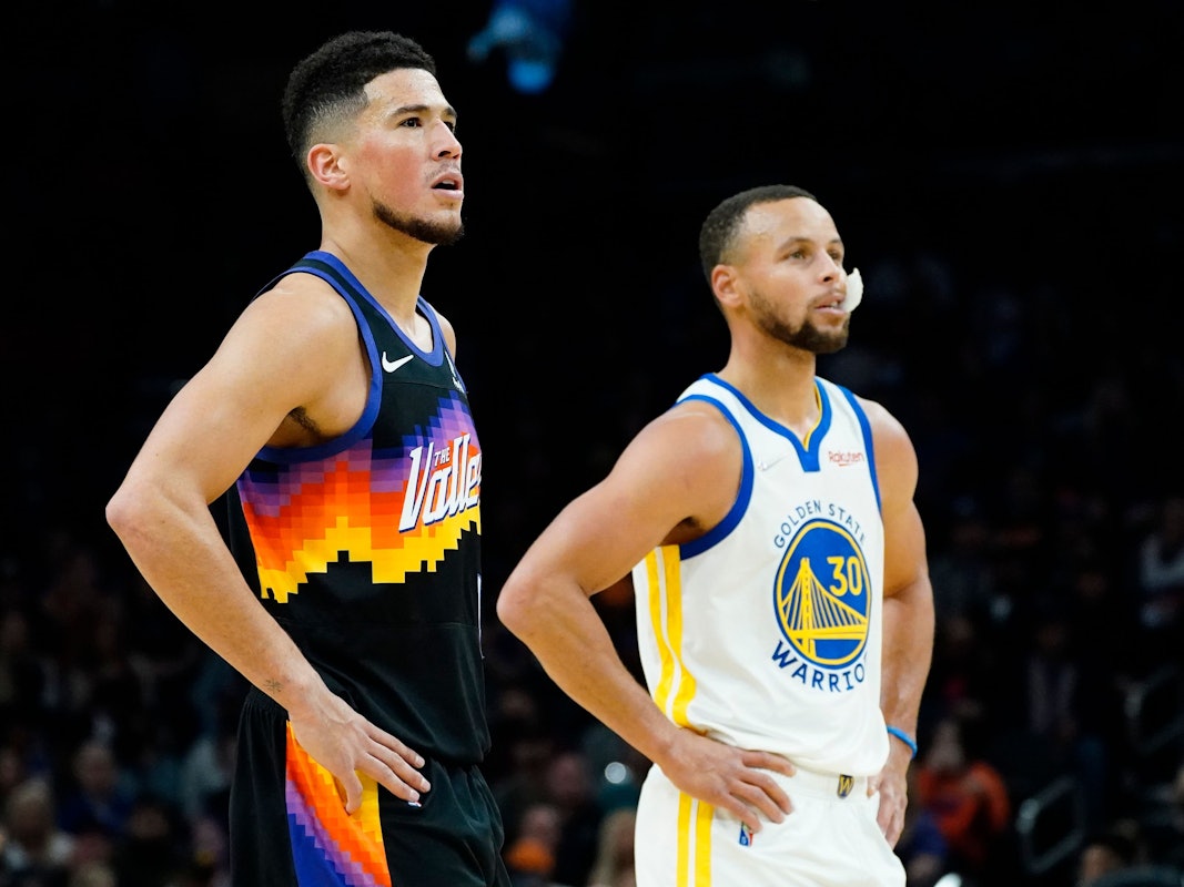 Suns-Guard Devin Booker (l.) und Warriors-Star Stephen Curry (r.) stützen jeweils ihre Arme in die Hüfte und schauen in Richtung Korb.