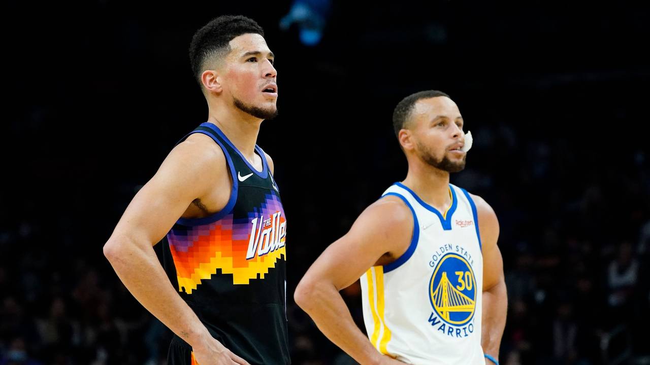 Suns-Guard Devin Booker (l.) und Warriors-Star Stephen Curry (r.) stützen jeweils ihre Arme in die Hüfte und schauen in Richtung Korb.