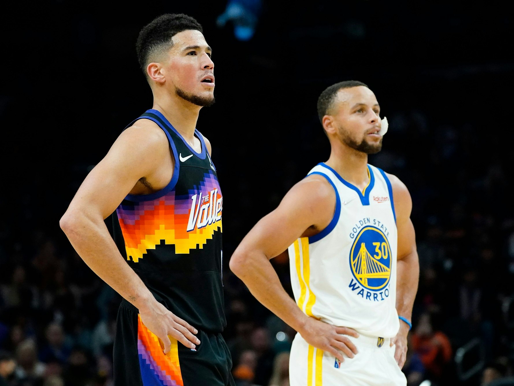 Suns-Guard Devin Booker (l.) und Warriors-Star Stephen Curry (r.) stützen jeweils ihre Arme in die Hüfte und schauen in Richtung Korb.