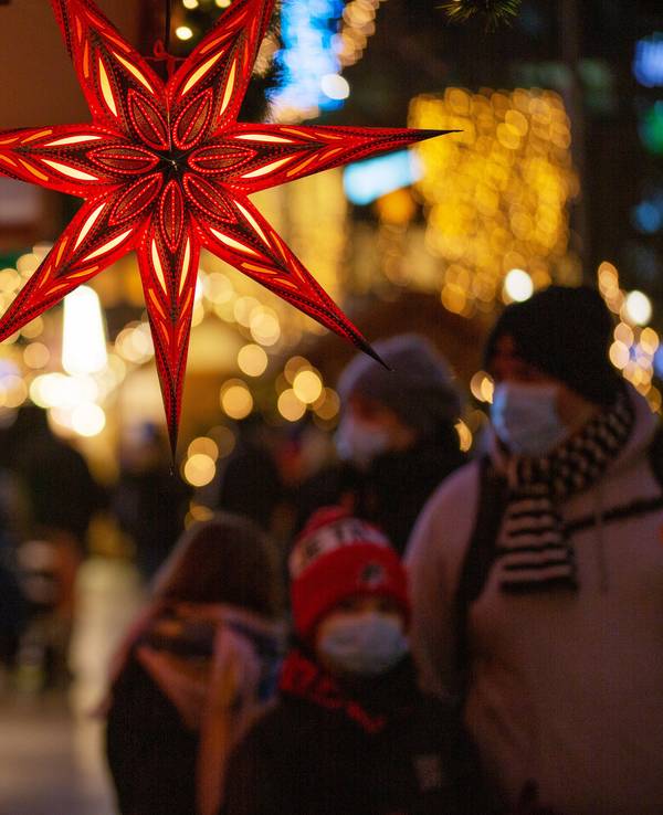 Eine Familie geht mit Mund-Nasen-Schutz auf einem Weihnachtsmarkt an einem Marktstand mit leuchtenden Sternen vorbei.