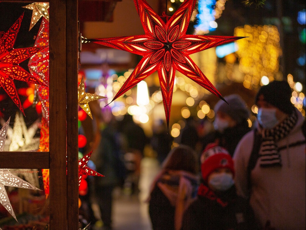 Eine Familie geht mit Mund-Nasen-Schutz auf einem Weihnachtsmarkt an einem Marktstand mit leuchtenden Sternen vorbei.