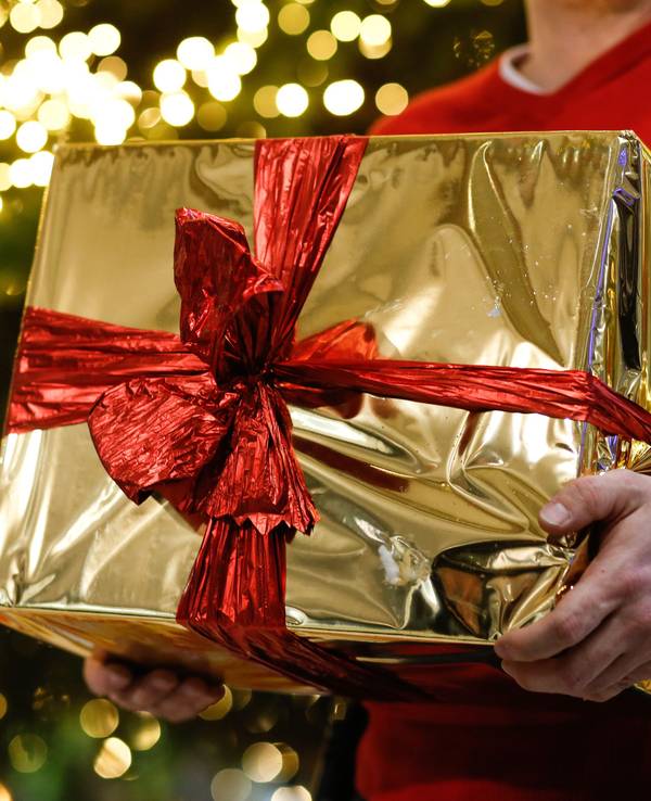 Ein Mann hält ein golden eingepacktes Weihnachtsgeschenk in den Händen. Im Hintergrund ist ein Weihnachtsbaum zu sehen.