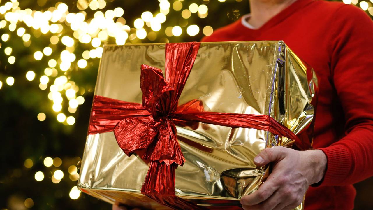 Ein Mann hält ein golden eingepacktes Weihnachtsgeschenk in den Händen. Im Hintergrund ist ein Weihnachtsbaum zu sehen.