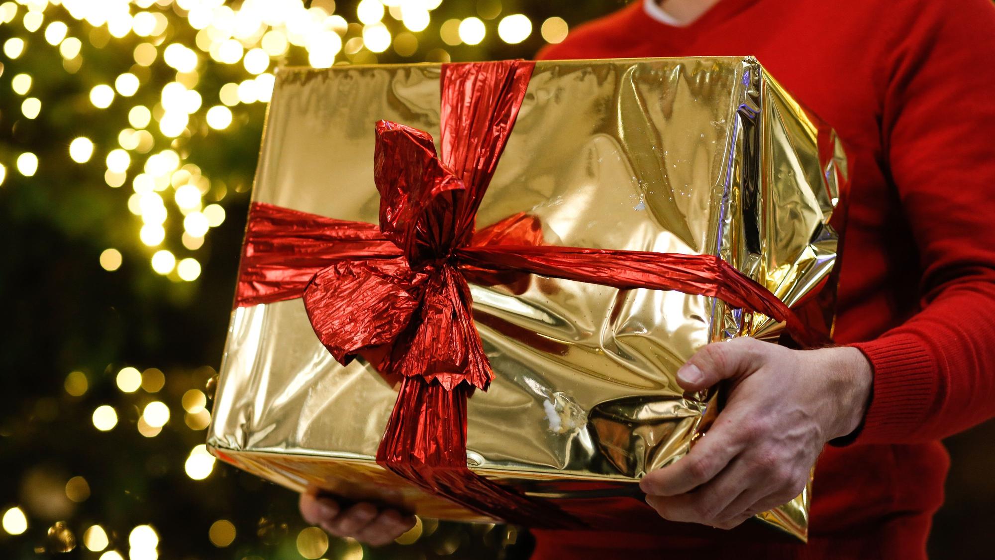 Ein Mann hält ein golden eingepacktes Weihnachtsgeschenk in den Händen. Im Hintergrund ist ein Weihnachtsbaum zu sehen.