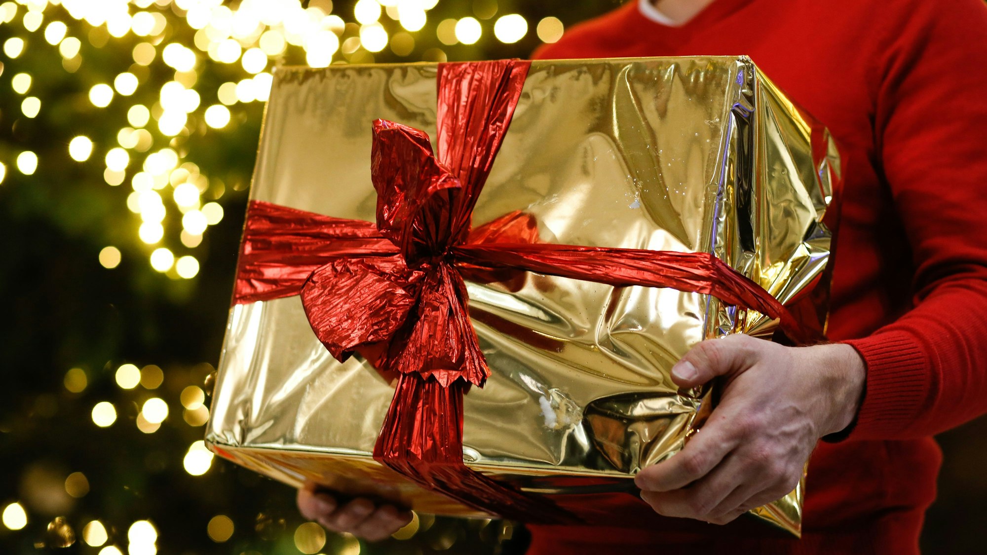 Ein Mann hält ein golden eingepacktes Weihnachtsgeschenk in den Händen. Im Hintergrund ist ein Weihnachtsbaum zu sehen.