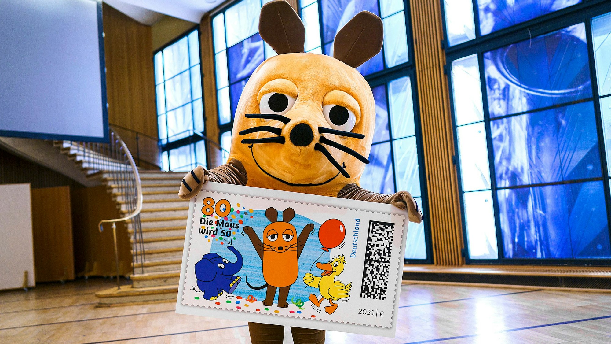 Die Hauptdarstellerin der Sendung mit der Maus hält eine überdimensionale Briefmarke mit ihren Darsteller-Kollegen Elefant und Ente in den Händen.