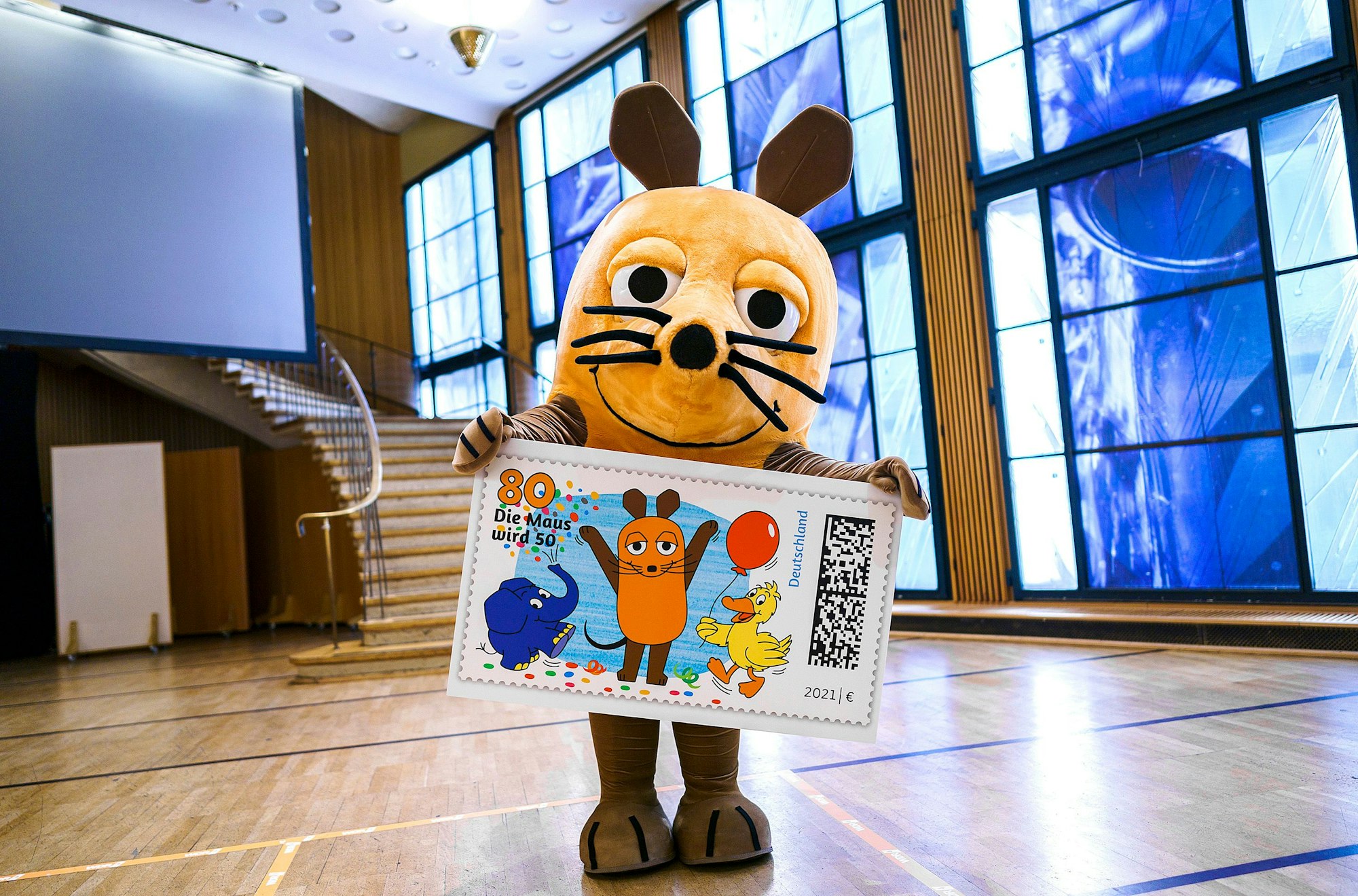 Die Hauptdarstellerin der Sendung mit der Maus hält eine überdimensionale Briefmarke mit ihren Darsteller-Kollegen Elefant und Ente in den Händen.