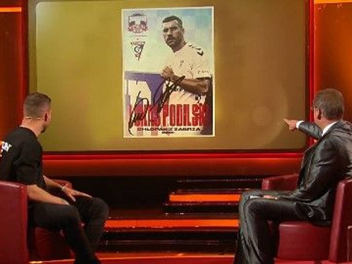 Lukas Podolski schaut mit Günther Jauch auf die Autogrammkarte.