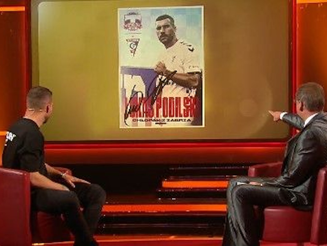 Lukas Podolski schaut mit Günther Jauch auf die Autogrammkarte.