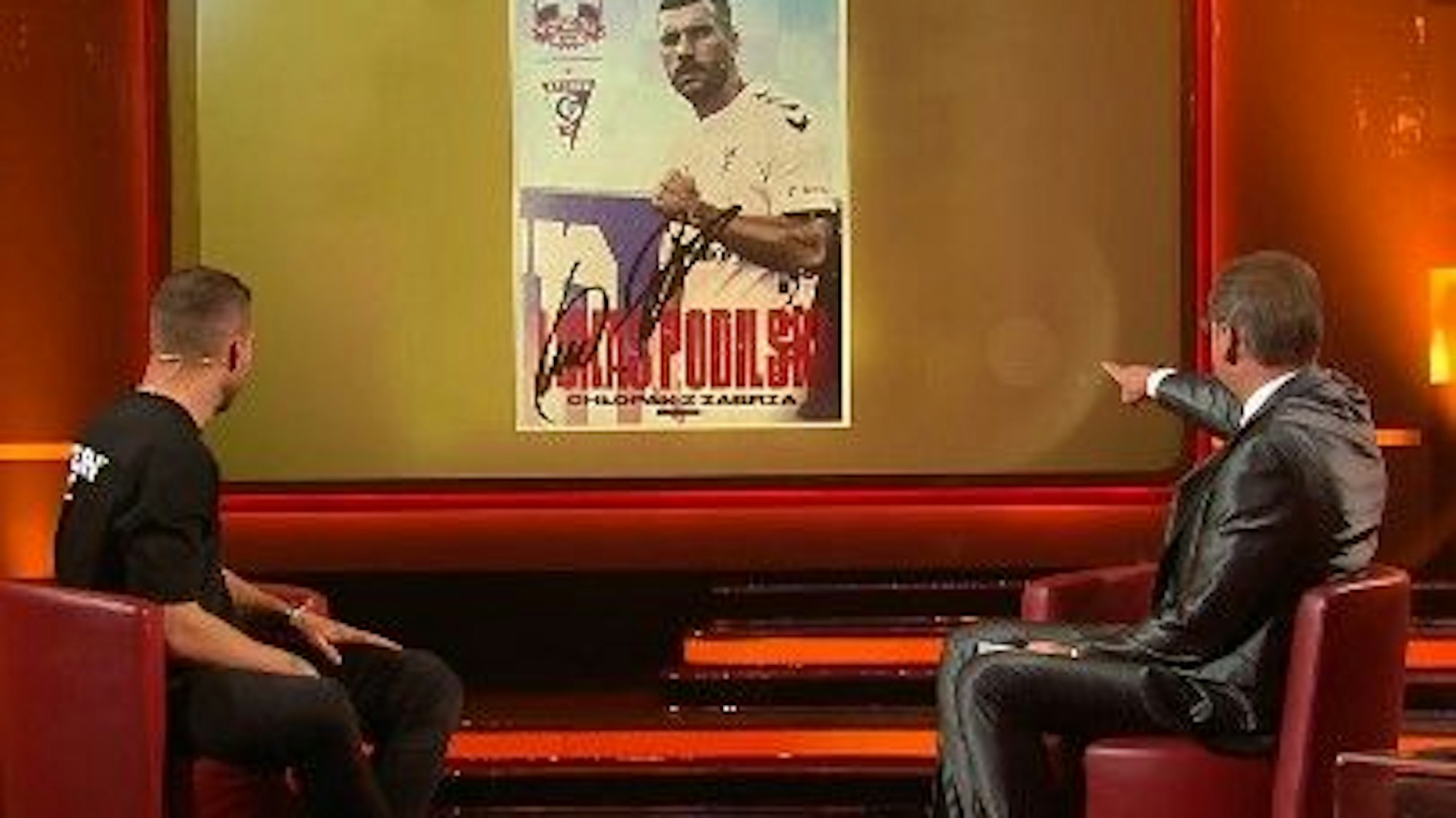 Lukas Podolski schaut mit Günther Jauch auf die Autogrammkarte.