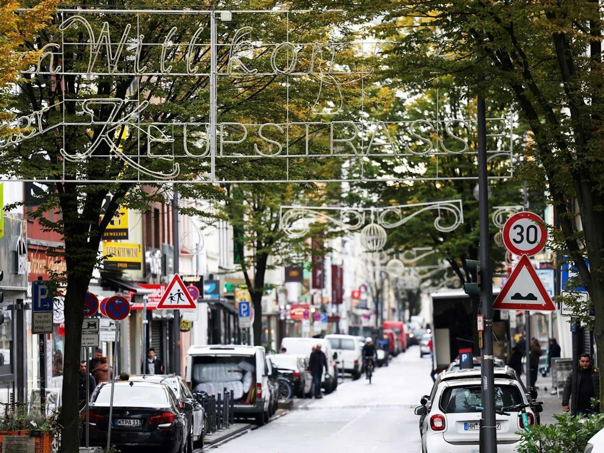 Köln: Die Keupstrasse mit mehreren Geschäften, Fußgängern und Autos.