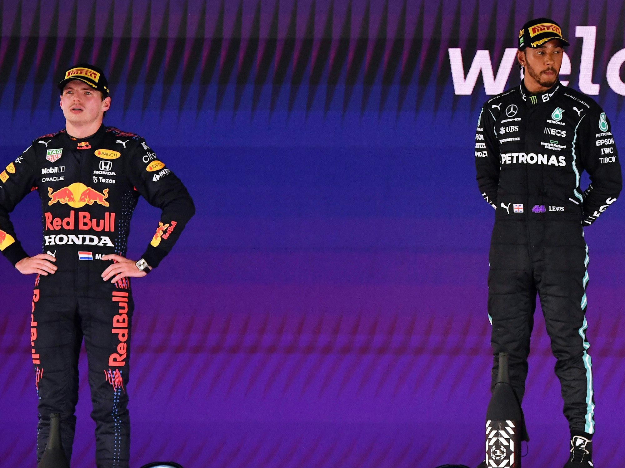 Max Verstappen steht links auf dem Podium, Lewis Hamilton rechts. Beide tragen Kappen. Verstappen hat die Hände in den Hüften.