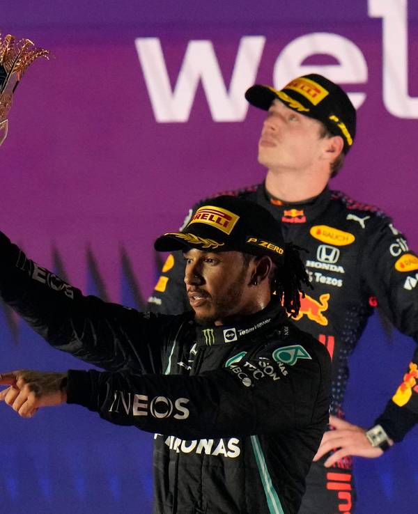 Lewis Hamilton jubelt nach einem Sieg in der Formel 1 mit Pokal, Max Verstappen steht zerknirscht dahinter.