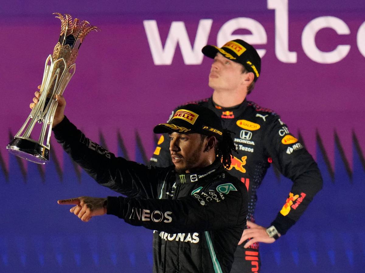 Lewis Hamilton jubelt nach einem Sieg in der Formel 1 mit Pokal, Max Verstappen steht zerknirscht dahinter.