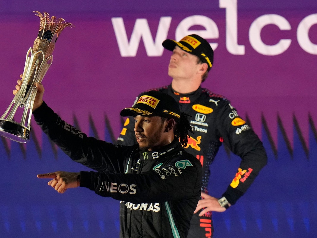 Lewis Hamilton jubelt nach einem Sieg in der Formel 1 mit Pokal, Max Verstappen steht zerknirscht dahinter.