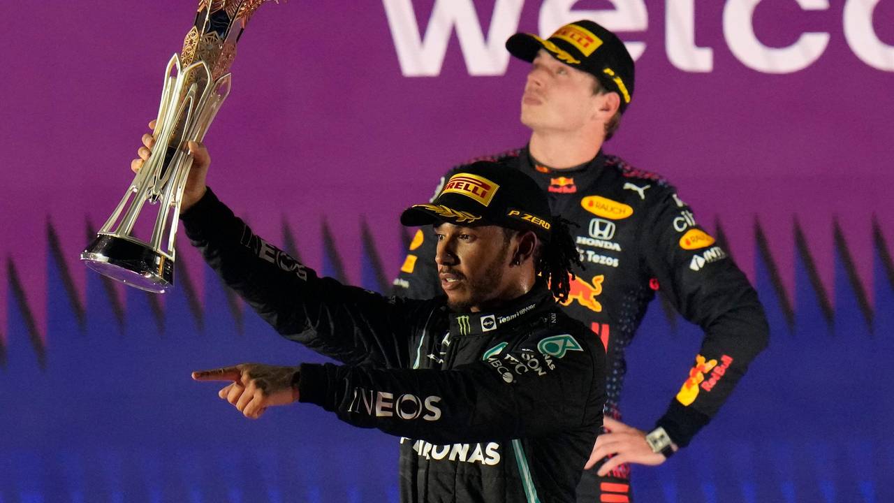 Lewis Hamilton jubelt nach einem Sieg in der Formel 1 mit Pokal, Max Verstappen steht zerknirscht dahinter.