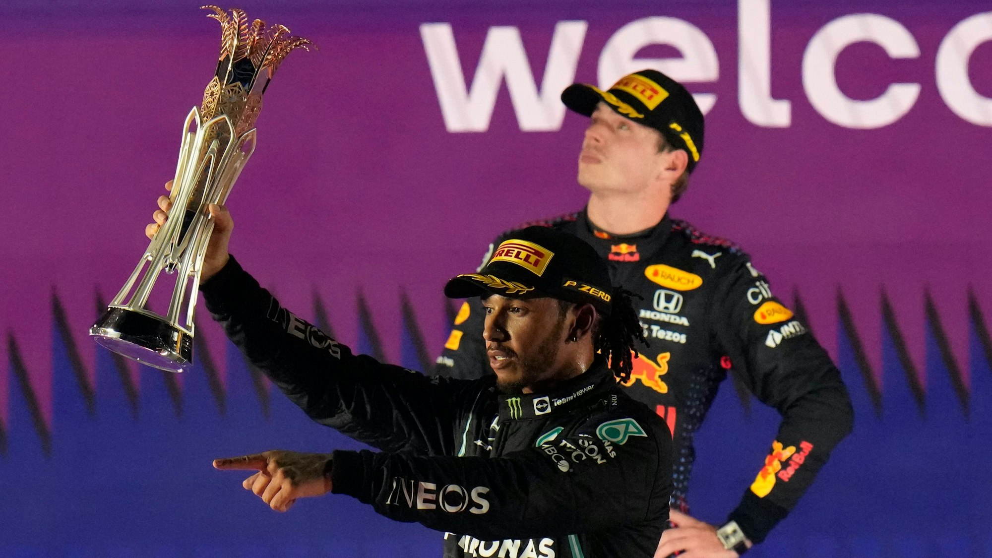 Lewis Hamilton jubelt nach einem Sieg in der Formel 1 mit Pokal, Max Verstappen steht zerknirscht dahinter.