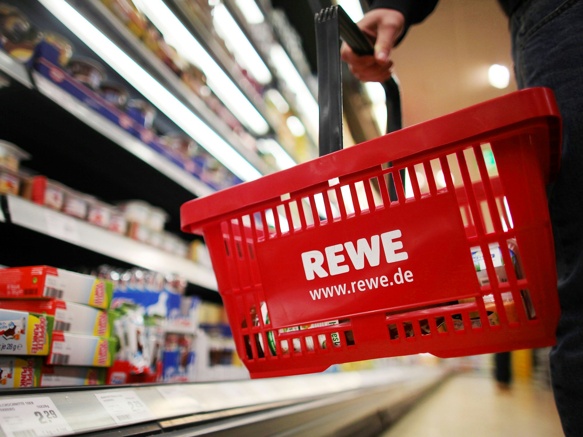 Ein Kunde steht mit einem roten Einkaufskorb mit der Aufschrift „Rewe“ vor einem Einkaufsregal.