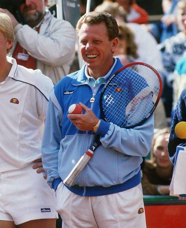 Boris Becker und Günther Bosch stehen auf dem Tennisplatz. Becker lehnt sich auf das Netz, Bosch hält einen Schläger und einen Ball.